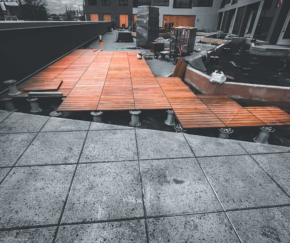 A rooftop deck with partially installed wooden planks, supported by pedestals, in the midst of construction with building materials nearby.