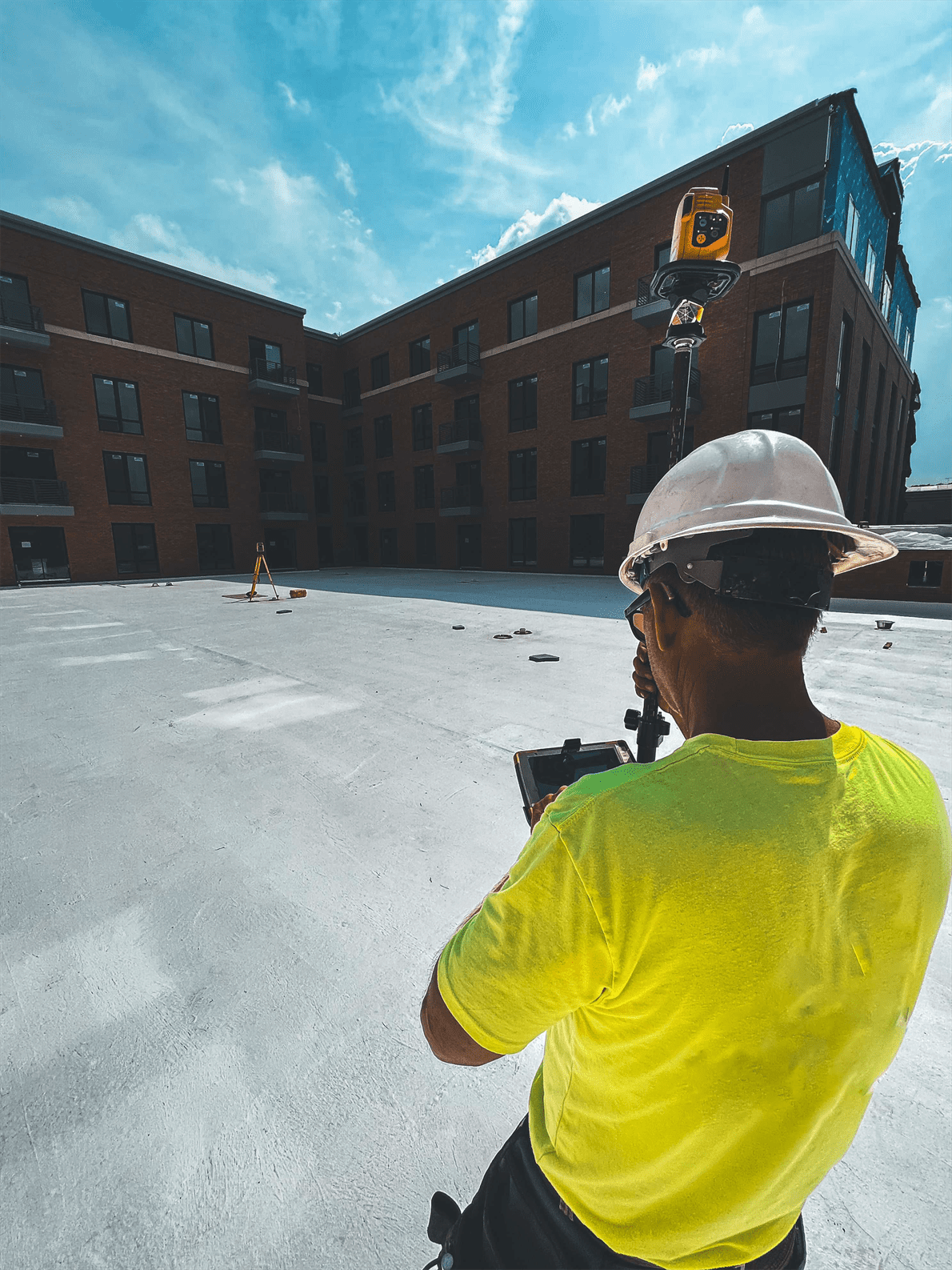 A construction worker in a neon shirt and hard hat uses a tablet to survey an empty rooftop space with apartment buildings in the background.